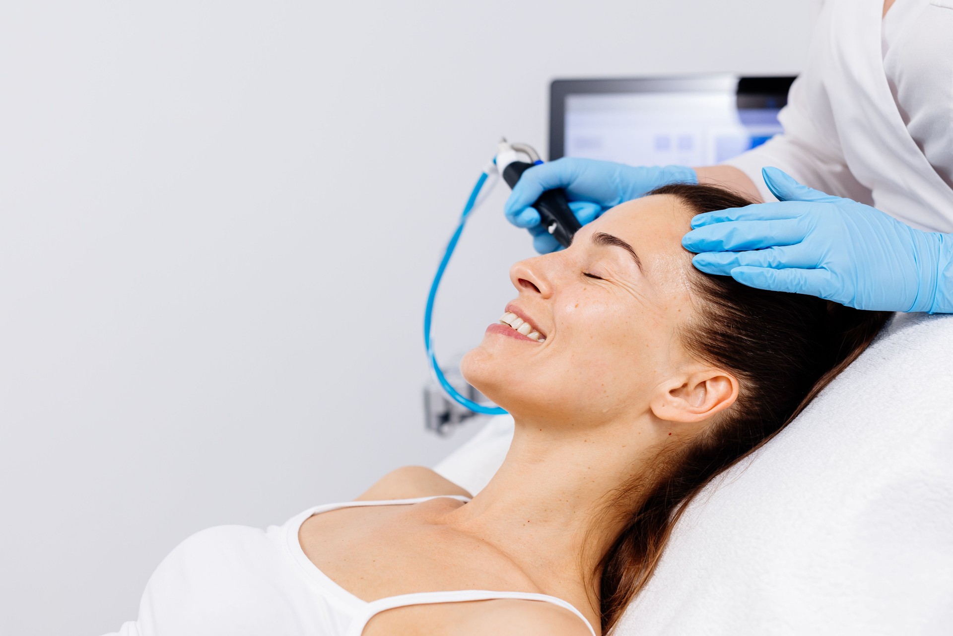 Una mujer de mediana edad recibe tratamiento facial en un salón de belleza profesional. Una cosmetóloga de cerca manipula un dispositivo de hidropeeling para limpiar y rejuvenecer la piel del rostro.